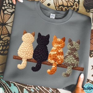 Könnte beinhalten: Graues Sweatshirt mit einem Design von vier Katzen, die auf einem Ast sitzen. Die Katzen sind in Creme, Schwarz, Orange und Grau dargestellt, mit strukturierter Fell. Das Design befindet sich mittig auf der Vorderseite des Sweatshirts.