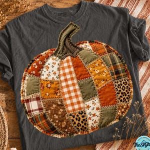May include: A charcoal gray t-shirt featuring a large, central pumpkin graphic composed of various fabric patches. The pumpkin design includes segments of orange, brown, green, white, and leopard print patterns, all joined by visible zigzag stitching.