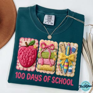 Pu&ograve; includere: Felpa Comfort Colors verde acqua con ricami a tema scolastico. Presenta una mela, libri e materiale scolastico. Il testo "100 DAYS OF SCHOOL" &egrave; ricamato sotto. Una collana d'oro &egrave; drappeggiata sulla camicia.