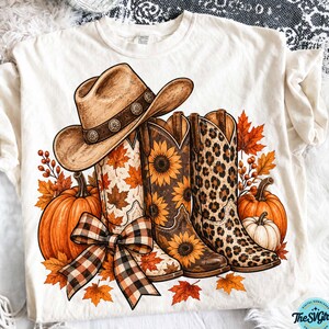 Puede incluir: Una camiseta color crema con un estampado gráfico de un sombrero de vaquero marrón, tres botas vaqueras con patrones de girasoles, hojas de arce y leopardo, calabazas naranjas y un lazo a cuadros, rodeado de hojas de otoño.