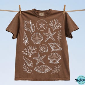 Puede incluir: Una camiseta marr&oacute;n con ilustraciones blancas de varias conchas marinas, incluyendo estrellas de mar, caracolas y coral. La camiseta est&aacute; colgada en un tendedero con pinzas de madera sobre un fondo azul claro. El dise&ntilde;o es una descarga digital.