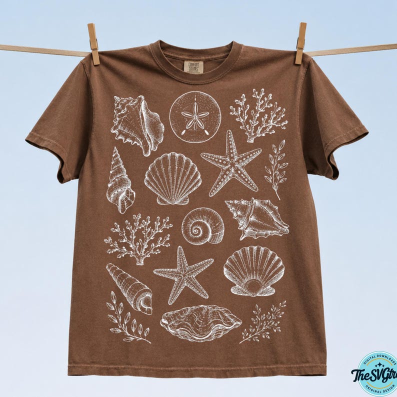 Puede incluir: Una camiseta marr&oacute;n con ilustraciones blancas de varias conchas marinas, incluyendo estrellas de mar, caracolas y coral. La camiseta est&aacute; colgada en un tendedero con pinzas de madera sobre un fondo azul claro. El dise&ntilde;o es una descarga digital.