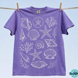 Puede incluir: Una camiseta morada con ilustraciones blancas de conchas marinas, incluyendo estrellas de mar, caracolas y coral. La camiseta est&aacute; colgada en un tendedero sobre un fondo azul claro. El dise&ntilde;o es una descarga digital de The SVG Girl.