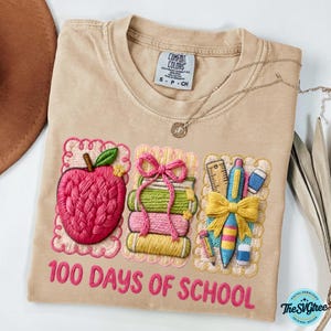 Pu&ograve; includere: T-shirt Comfort Colors color cachi con ricami a tema scolastico. Presenta una mela rosa, libri impilati e materiale scolastico. Il testo "100 DAYS OF SCHOOL" &egrave; ricamato in rosa.