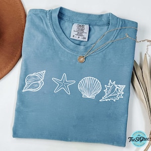 May include: A blue t-shirt with white seashell graphics, including a conch, starfish, and scallop shell. The shirt has a Comfort Colors tag and a gold necklace. The design is from The SVGtree.