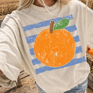 Könnte beinhalten: Cremefarbenes T-Shirt mit einer großen orangefarbenen Fruchtgrafik, einem braunen Stiel und einem grünen Blatt, vor blauen horizontalen Streifen. Das Design hat ein strukturiertes, handgezeichnetes Aussehen und erzeugt eine lässige, künstlerische Ästhetik.