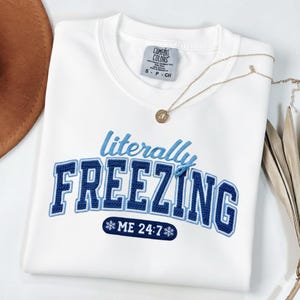 May include: White sweatshirt with the phrase "literally FREEZING" embroidered in blue. The sweatshirt has a crew neck and a tag that says "Comfort Colors". A gold necklace with a pendant is draped across the front.