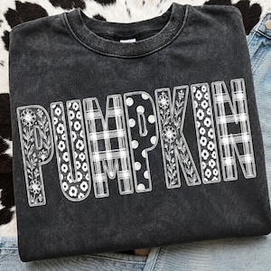 May include: A charcoal gray acid-wash sweatshirt featuring the word PUMPKIN printed in large white letters. Each letter contains a unique pattern, including floral designs, a plaid check, and polka dots, all outlined in a thin white border.