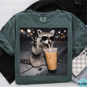 May include: Dark green long-sleeve shirt featuring a graphic of a raccoon wearing sunglasses and drinking an iced coffee. The raccoon is giving a peace sign. The image has a nighttime city background. The shirt has a relaxed fit.