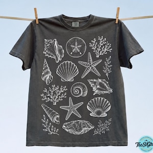 Puede incluir: Camiseta gris oscuro con ilustraciones blancas de conchas marinas, estrellas de mar y corales. La camiseta est&aacute; colgada en un tendedero con pinzas de madera. El dise&ntilde;o tiene un tema costero o de playa, ideal para el verano.