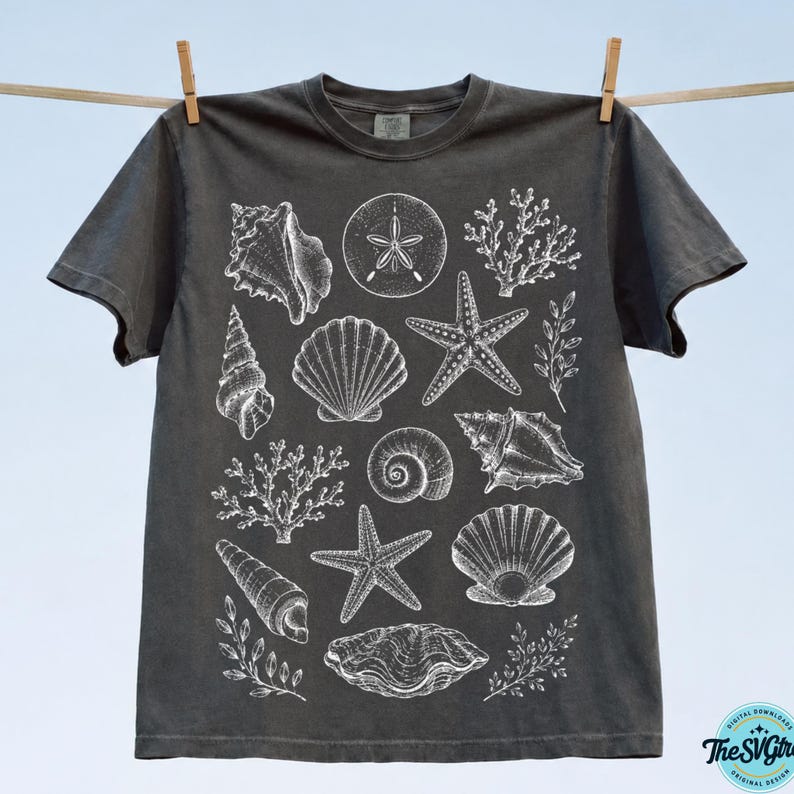 Puede incluir: Camiseta gris oscuro con ilustraciones blancas de conchas marinas, estrellas de mar y corales. La camiseta est&aacute; colgada en un tendedero con pinzas de madera. El dise&ntilde;o tiene un tema costero o de playa, ideal para el verano.