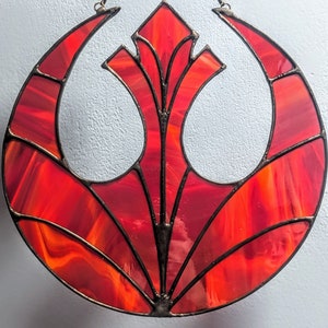 Peut inclure: Une fenêtre en vitrail représentant le symbole de l'Alliance rebelle de Star Wars. Le symbole est rouge et orange avec des lignes noires et est placé sur un fond blanc.