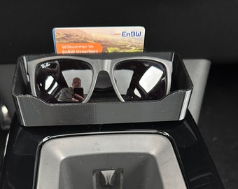 Puede incluir: Un portagafas de plástico negro con un par de gafas de sol negras en su interior. El portagafas está sujeto a la consola central de un coche. Una tarjeta con el texto "Willkommen im EnBW HyperNetz" es visible en el portagafas.