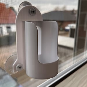 Könnte beinhalten: Weißer Plastikhalter mit Saugnäpfen, der an einem Fenster befestigt ist. Der Halter hat einen Schlitz für ein Telefon oder ein anderes Gerät.