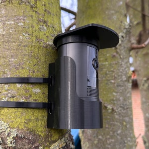 May include: A black security camera mounted on a tree trunk with black zip ties. The camera has a protective black visor and is designed for outdoor use. The tree bark is covered in green and gray lichen.
