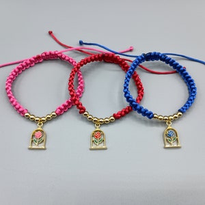 May include: Three braided friendship bracelets with gold-toned charms featuring a rose in a glass dome. The bracelets are pink, red, and blue.