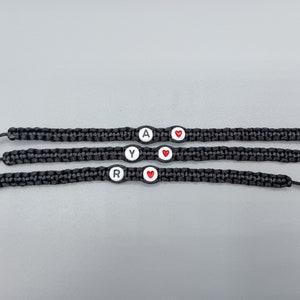 May include: Three black friendship bracelets with white beads and red heart beads. The beads are spelled "RAY" with a heart between each letter.