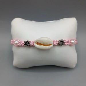 May include: A pink braided bracelet with a white seashell charm and silver flower accents.