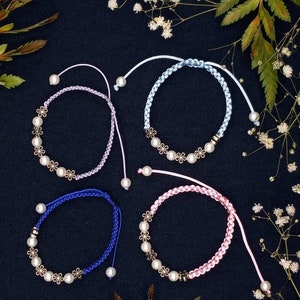 May include: Four handmade friendship bracelets with pearl and flower charms. The bracelets are in shades of pink, blue, purple, and light blue.