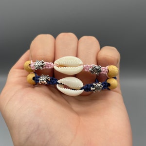 May include: Two bracelets with blue and pink cords, wooden beads, and silver turtle charms. Each bracelet features a white seashell charm.