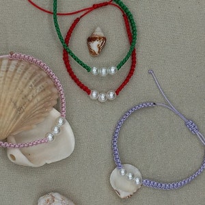 May include: Three bracelets with pearl accents. The bracelets are made with red, green, and purple string. The bracelets are displayed on a beige surface with seashells.