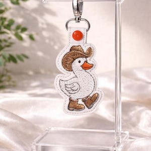 May include: A white embroidered keychain featuring a cartoon duck wearing a brown cowboy hat and boots. The duck has an orange beak and is attached to a silver keyring with a red accent.