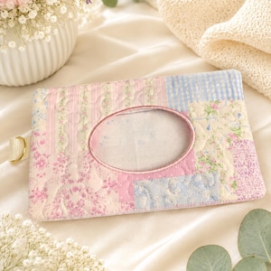 May include: A rectangular quilted fabric tissue holder featuring a patchwork design of pastel pink, blue, and cream floral, striped, and gingham patterns. The case has a central oval opening with a finished edge and a small side loop with a gold ring.