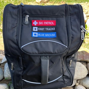 May include: Black ski bag with a white zipper and a white stripe around the edge. The bag has three patches on the front: "Ski Patrol", "First Tracks", and "Blue Grouse".