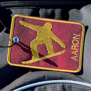 Puede incluir: Un parche de tela color granate con costuras doradas y una silueta dorada de una persona haciendo snowboard. El texto "AARON" está bordado en dorado en el lado derecho del parche.