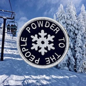 May include: A circular embroidered patch with a black background and a white border. The center features a white snowflake icon surrounded by the white text "POWDER TO THE PEOPLE" arranged in a circular pattern around the snowflake.