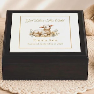 May include: A dark brown wooden keepsake box with a square white tile insert on the lid. The tile features a gold-bordered illustration of a lamb, the text "God Bless This Child," the name "Emma Ann," and "Baptized September 7, 2025."