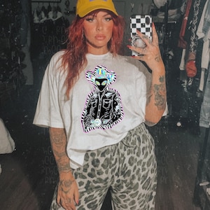 Könnte beinhalten: Weißes T-Shirt mit einer Grafik eines Außerirdischen, der einen Cowboyhut und eine Jacke trägt, umrandet von einem Zebrastreifenmuster. Der Außerirdische hat große schwarze Augen und eine Gürtelschnalle. Das Shirt wird mit einer Hose mit Leopardenmuster getragen.