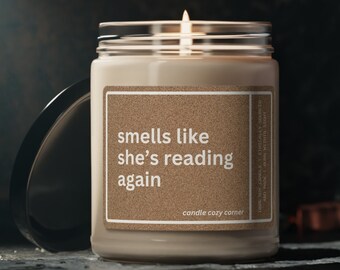 May include: A lit, beige-colored candle in a glass jar with a silver lid. The label reads "smells like she's reading again" in white text. The candle is on a dark, textured surface.