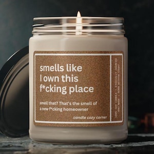 May include: A lit, cream-colored candle in a glass jar with a black lid. The label reads "smells like I own this f*cking place." The text below says, "smell that? That's the smell of a new f*cking homeowner."