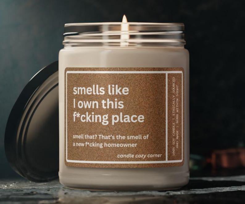May include: A lit, cream-colored candle in a glass jar with a black lid. The label reads "smells like I own this f*cking place." The text below says, "smell that? That's the smell of a new f*cking homeowner."