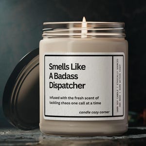 May include: A lit soy candle in a glass jar with a silver lid. The label reads "Smells Like A Badass Dispatcher." The candle is infused with a fresh scent. The text "candle cozy corner" is at the bottom of the label.
