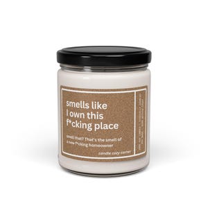 May include: A white candle in a glass jar with a black lid. The label reads "smells like I own this f*cking place." Additional text says "smell that? That's the smell of a new f*cking homeowner." The candle is from Candle Cozy Corner.