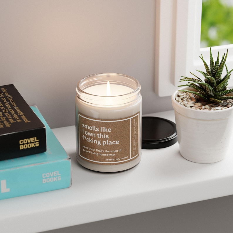 May include: A lit, scented candle in a clear glass jar with a brown label that reads "Smells like I own this f*cking place." The candle sits on a white shelf next to a small potted succulent and two books.