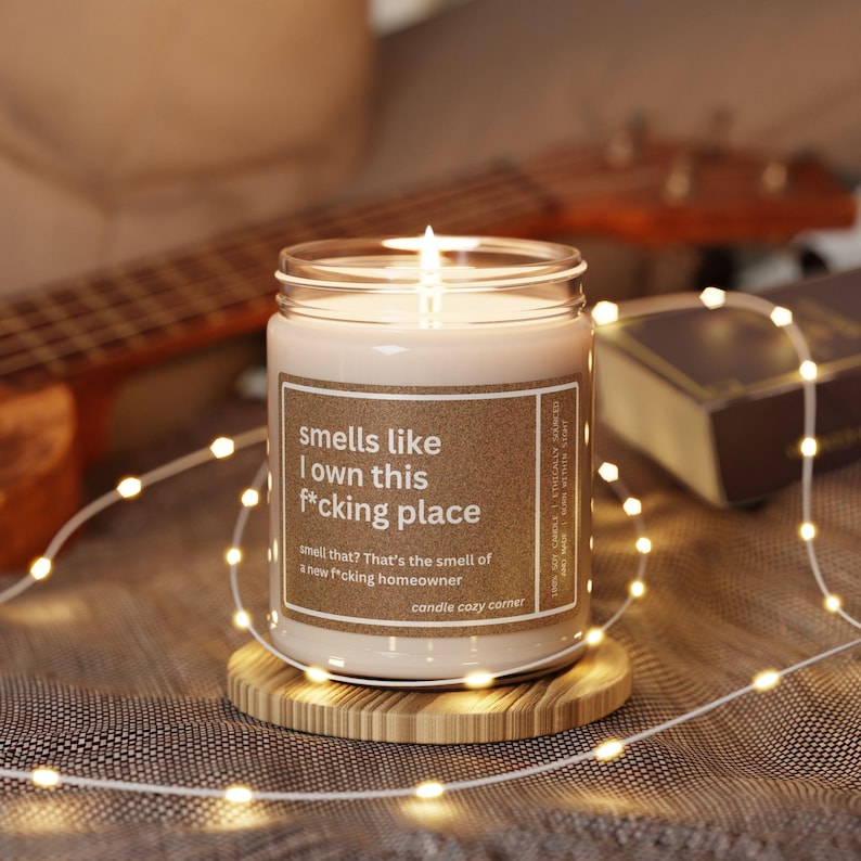 May include: A lit candle in a glass jar with a brown label that reads "smells like I own this f*cking place." The candle sits on a wooden coaster, surrounded by string lights. A guitar and a book are in the background.