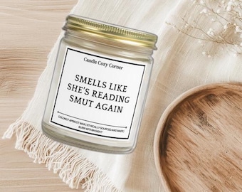 May include: A clear glass jar candle with a gold lid, featuring a white label with black text that reads "SMELLS LIKE SHE'S READING SMUT AGAIN." The candle is on a white cloth with a wooden plate nearby. The text "Candle Cozy Corner" is at the top of the label.
