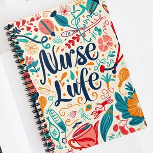 May include: Spiral-bound notebook with a cream-colored cover featuring the words "Nurse Life" in navy blue script. The cover is decorated with colorful illustrations of medical tools, flowers, and other decorative elements.
