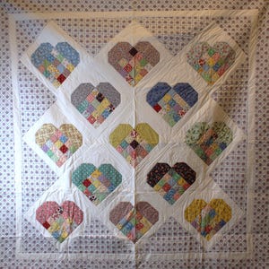 Op de afbeelding: Een handgemaakte quilt met een herhalend hartpatroon. Elk hart is samengesteld uit verschillende stofresten in een patchwork-ontwerp. De quilt heeft een witte achtergrond met een patroonrand, wat een vintage esthetiek laat zien.