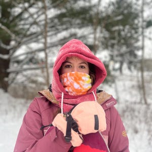 Może przedstawiać: Osoba w różowej kurtce z kapturem i maską na twarz w stylu tie-dye trzyma deskę snowboardową. Osoba stoi w zasneżonym lesie.