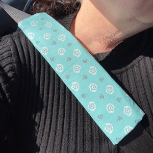 May include: A turquoise blue car seatbelt cover with a white floral pattern. The cover is made of fabric and has a soft texture.