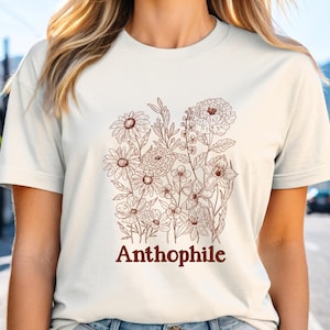 May include: Cream-colored t-shirt featuring a brown floral illustration with various flower types and foliage. The word "Anthophile" is printed in brown script below the floral design. The shirt has short sleeves and a crew neck.