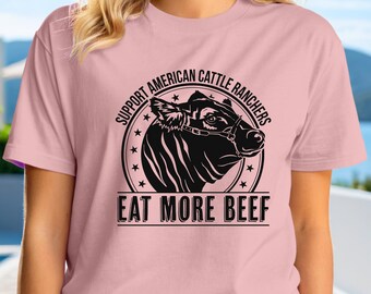 Puede incluir: Camiseta rosa claro con un gráfico negro de una cabeza de vaca dentro de un círculo. El texto dice "SUPPORT AMERICAN CATTLE RANCHERS" y "EAT MORE BEEF". La camiseta tiene mangas cortas y cuello redondo.