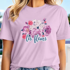 May include: A light purple t-shirt featuring a floral design with pink, purple, and white flowers. The words "Les Fleurs" are written in a cursive font below the flowers. The shirt is made of a soft material.