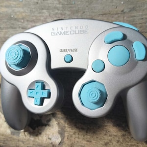 May include: A silver Nintendo GameCube controller featuring light blue buttons, a light blue D-pad, and two light blue analog sticks. The controller has the Nintendo GameCube logo and the word START/PAUSE printed in black on the front face.