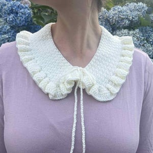 Puede incluir: Cuello de crochet color crema con borde con volantes y cierre de lazo. El cuello se lleva sobre una camisa de manga larga de color lila claro. Las flores de hortensia azules están en el fondo.