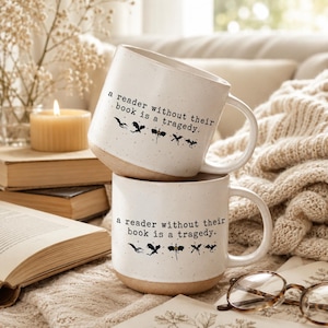 Puede incluir: Dos tazas de cerámica blanca con acabado moteado y base color canela. Cada taza presenta el texto negro 'a reader without their book is a tragedy.' seguido de una fila de pequeñas siluetas de dragones negras. Tienen un asa y borde curvos.